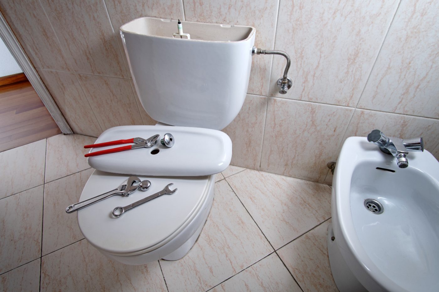Bathroom with toilet seat open displaying wrenches and pliers, cistern tank above, and bidet on right side